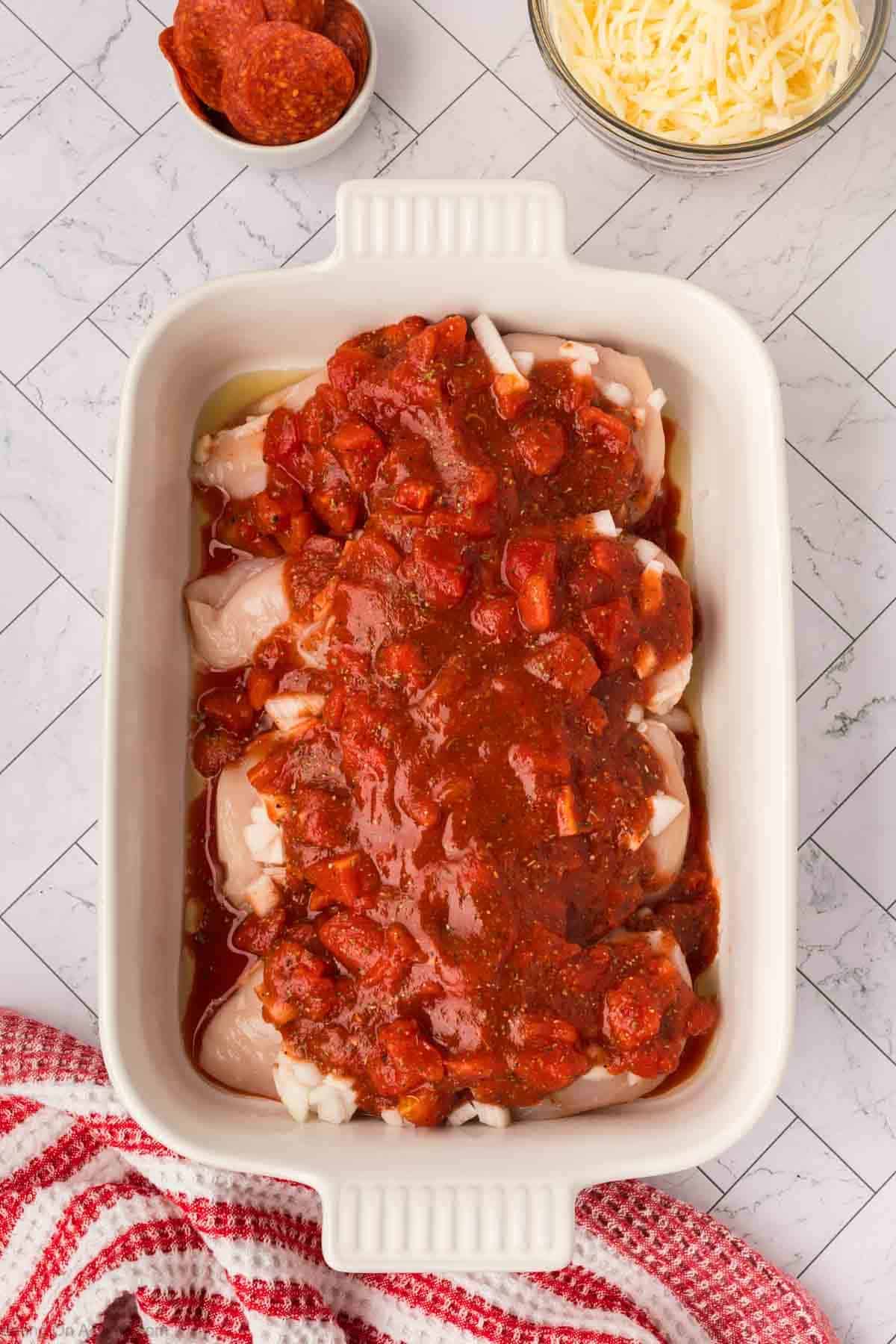 A white baking dish filled with raw chicken breasts topped with tomato sauce and chopped tomatoes sits nearby bowls of shredded cheese and pepperoni slices—perfect ingredients for a Low Carb Pizza Chicken Recipe on a tiled surface with a red and white cloth.