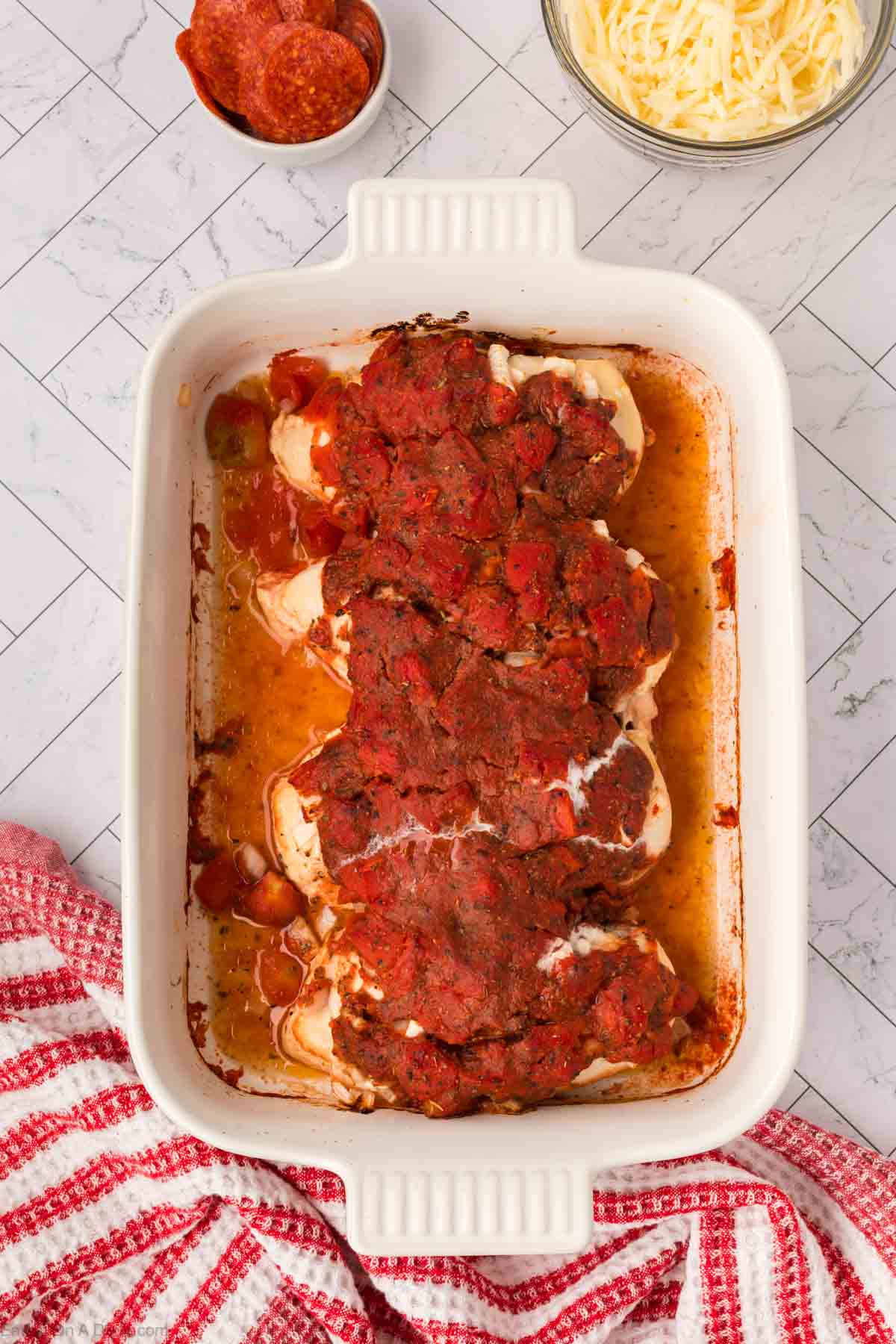 A white baking dish filled with Low Carb Pizza Chicken Recipe—baked chicken breasts topped with tomato sauce—sits on a white tiled surface. Nearby are a bowl of pepperoni slices, shredded cheese, and a red patterned towel.