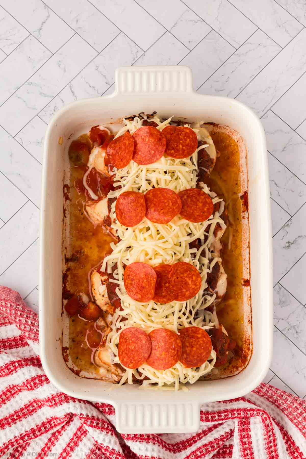 A white baking dish holds cooked chicken breasts topped with marinara sauce, shredded mozzarella, and pepperoni—this Low Carb Pizza Chicken Recipe sits on a marble surface beside a red and white checkered towel.