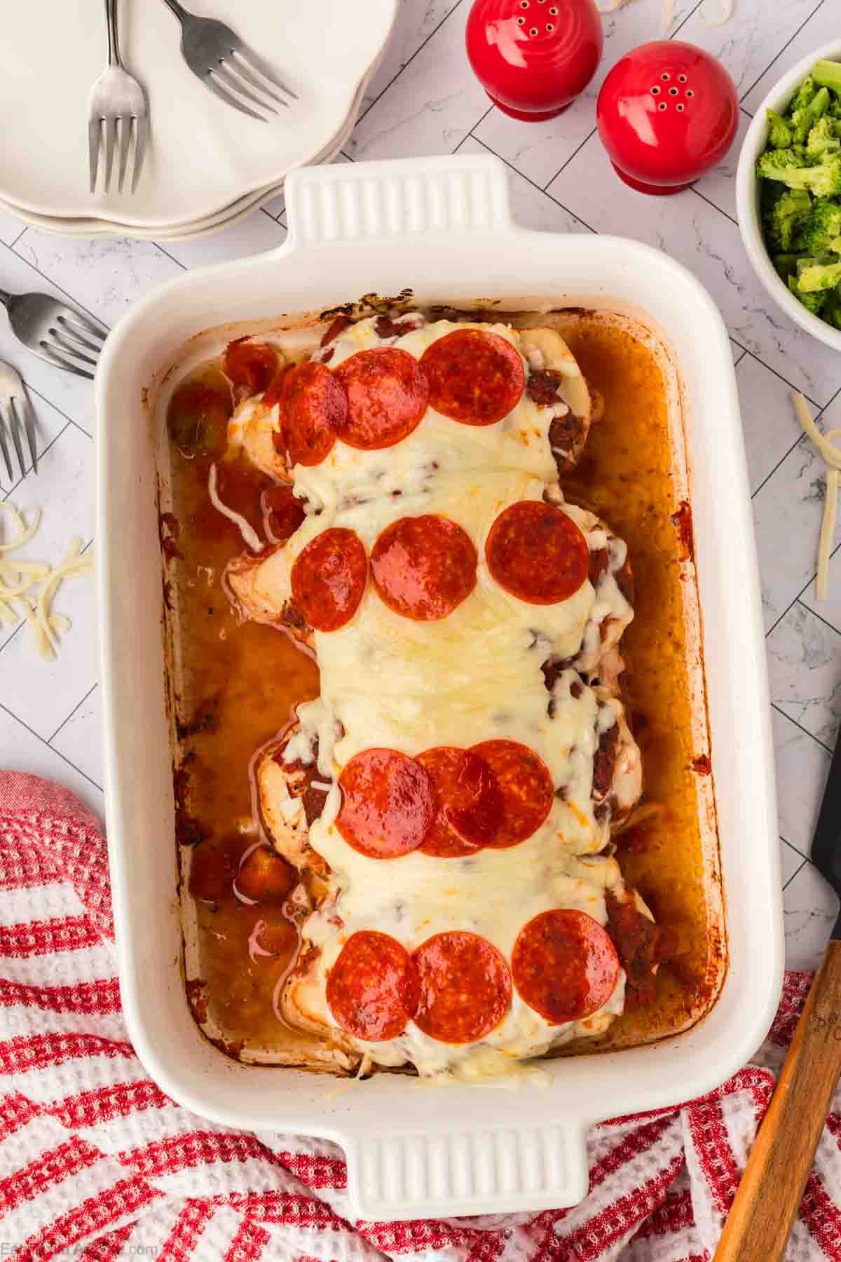 A white baking dish filled with Low Carb Pizza Chicken Recipe—chicken breasts topped with melted cheese and pepperoni slices—set on a table with forks, a red-striped towel, broccoli, and red salt and pepper shakers nearby.