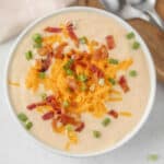 A bowl of creamy crockpot cauliflower soup recipe topped with shredded cheddar cheese, crispy bacon pieces, and chopped green onions, with two spoons on a wooden board in the background.