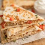 Three stacked chicken quesadilla wedges with melted cheese and diced tomatoes, garnished with chopped herbs on parchment paper. Inspired by a crockpot chicken quesadilla recipe, a bowl of sour cream is blurred in the background.
