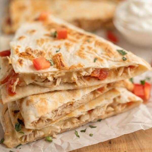 Three stacked chicken quesadilla wedges with melted cheese and diced tomatoes, garnished with chopped herbs on parchment paper. Inspired by a crockpot chicken quesadilla recipe, a bowl of sour cream is blurred in the background.