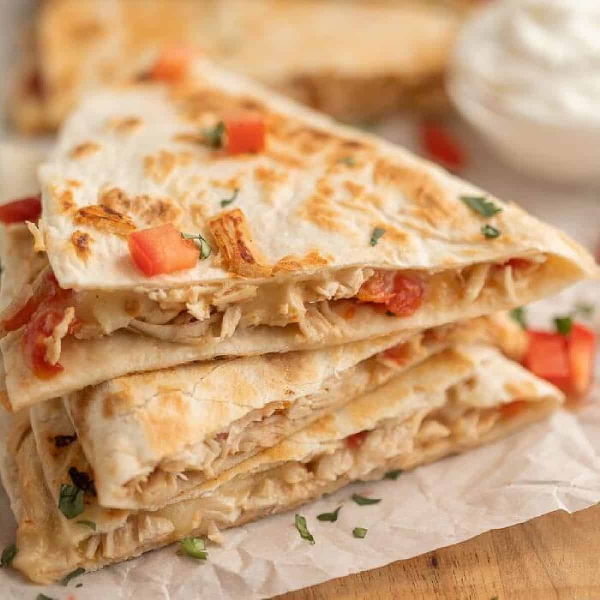 Three stacked chicken quesadilla wedges with melted cheese and diced tomatoes, garnished with chopped herbs on parchment paper. Inspired by a crockpot chicken quesadilla recipe, a bowl of sour cream is blurred in the background.