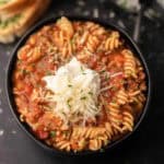 A black bowl filled with Gluten Free Crock Pot Lasagna Soup featuring rotini pasta in a tomato-based meat sauce, topped with shredded cheese, a dollop of ricotta, and chopped herbs. Slices of bread are visible in the background.