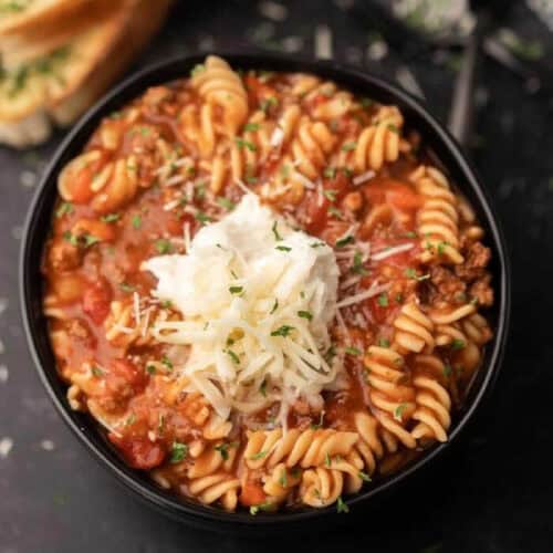 A black bowl filled with Gluten Free Crock Pot Lasagna Soup featuring rotini pasta in a tomato-based meat sauce, topped with shredded cheese, a dollop of ricotta, and chopped herbs. Slices of bread are visible in the background.