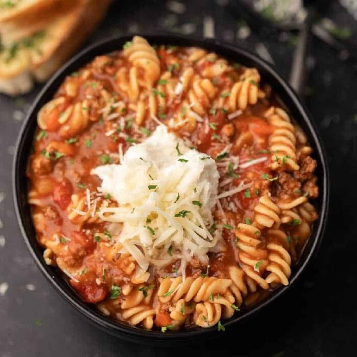 A black bowl filled with Gluten Free Crock Pot Lasagna Soup featuring rotini pasta in a tomato-based meat sauce, topped with shredded cheese, a dollop of ricotta, and chopped herbs. Slices of bread are visible in the background.