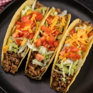Three hard shell tacos filled with seasoned ground beef made from our Easy Crockpot Taco Meat Recipe, shredded lettuce, diced tomatoes, and cheddar cheese, served on a black plate.