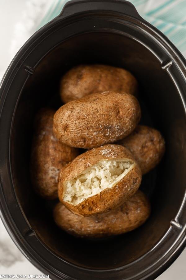 Multiple baked potatoes in a black crock pot with one cut opened 