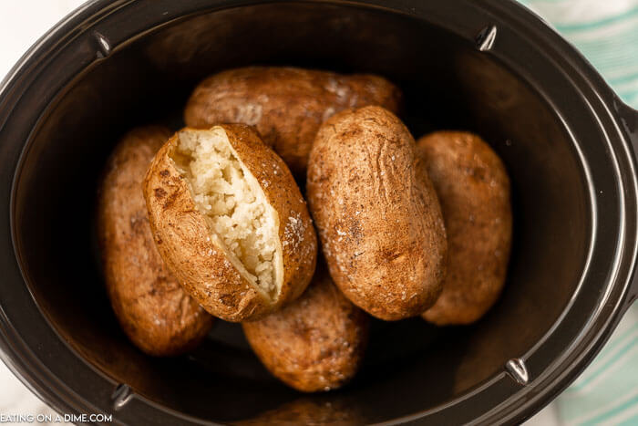 Multiple baked potatoes in a black crock pot with one cut opened 