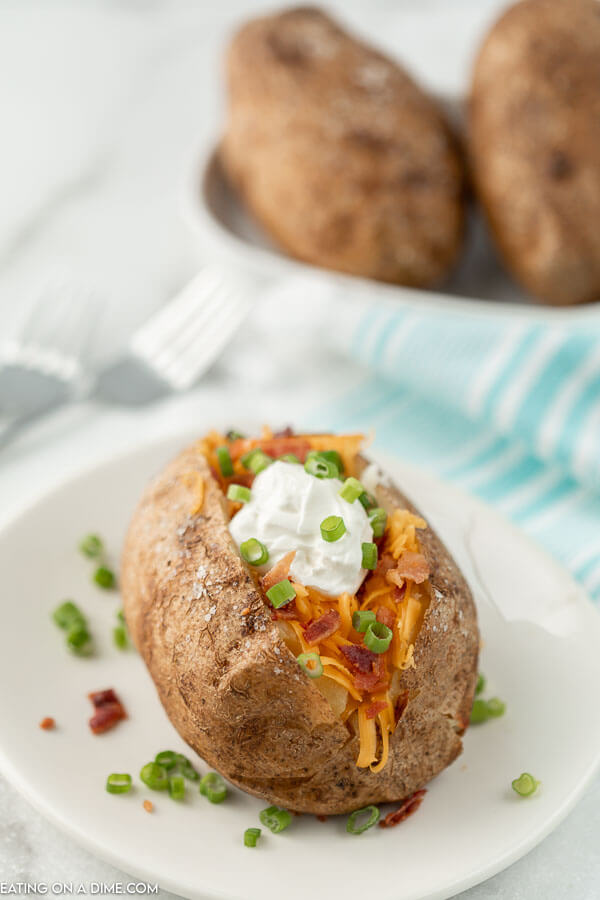 A baked potato on a white plate topped with shredded cheddar cheese, bacon bits, sour cream and chives with more potatoes behind it. 