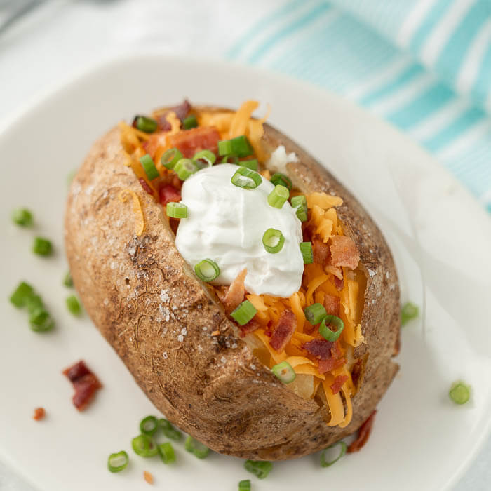 Close up of a baked potato on a white plate topped with shredded cheddar cheese, bacon bits, sour cream and chives.  