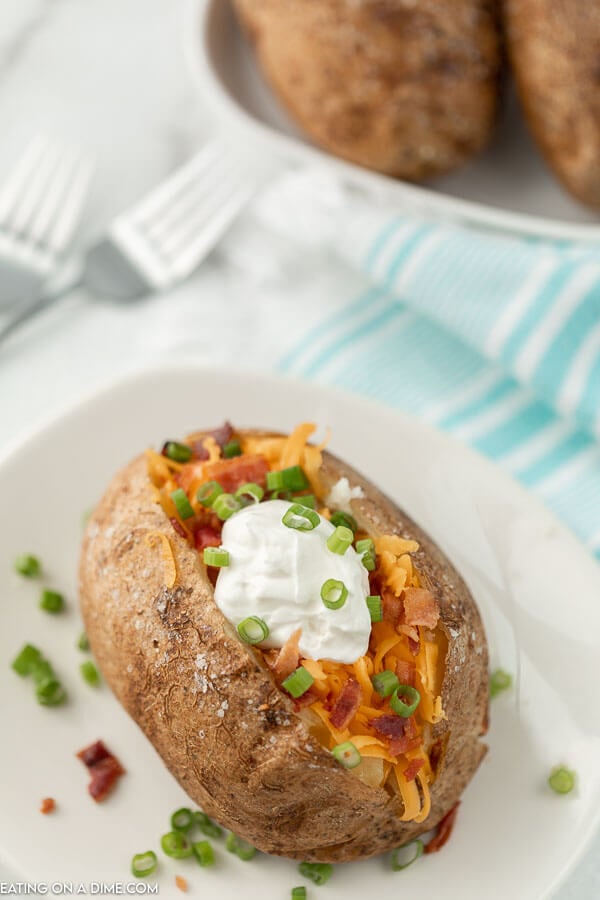 A baked potato on a white plate topped with shredded cheddar cheese, bacon bits, sour cream and chives.  