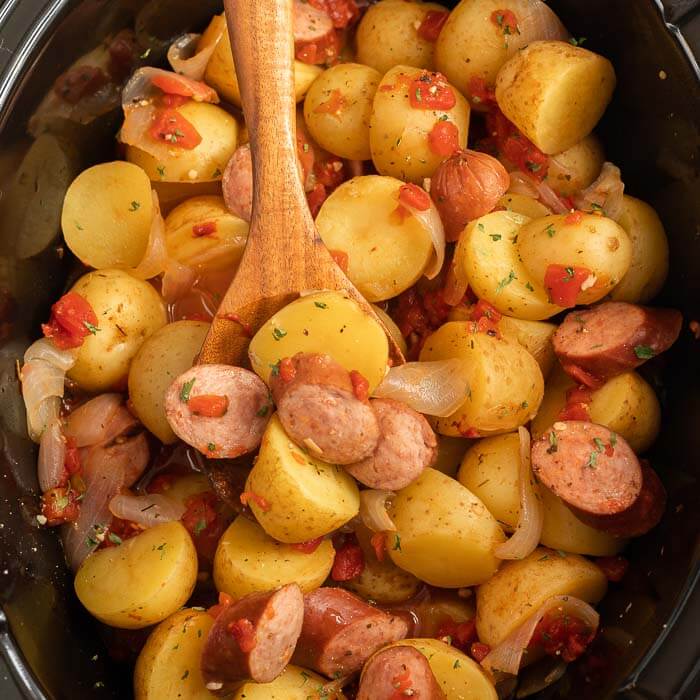 Close up of crock Pot Sausage and Potatoes in a black crock pot with a wooden spoon in the food in the crock pot.  