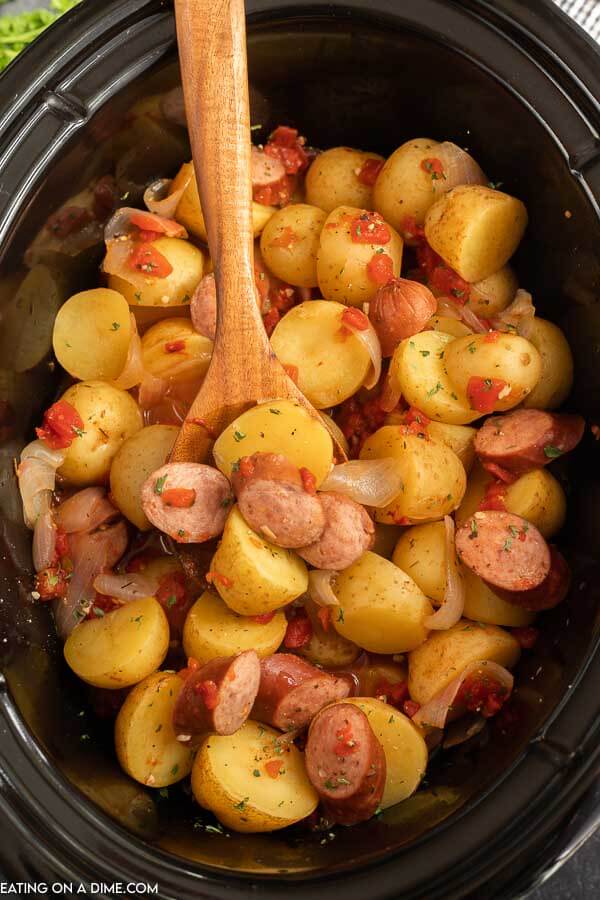 Crock Pot Sausage and Potatoes in a black crock pot with a wooden spoon in the food in the crock pot.  