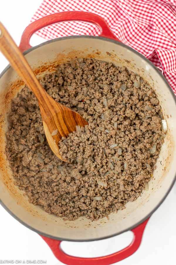 Close up image of ground beef being cooked in a dutch oven with a wooden spoon. 