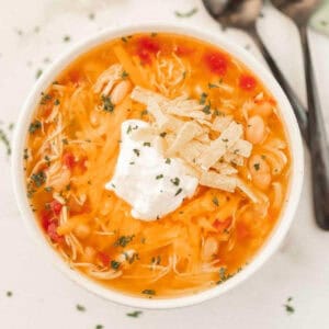 A bowl of creamy instant pot chicken taco soup topped with shredded cheese, a dollop of sour cream, crispy tortilla strips, and chopped herbs. Two spoons rest beside the bowl on a light surface.
