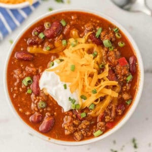 A bowl of Instant Pot Cowboy Chili topped with shredded cheddar cheese, a dollop of sour cream, and chopped green onions, featuring red beans and ground meat in a thick, red sauce.