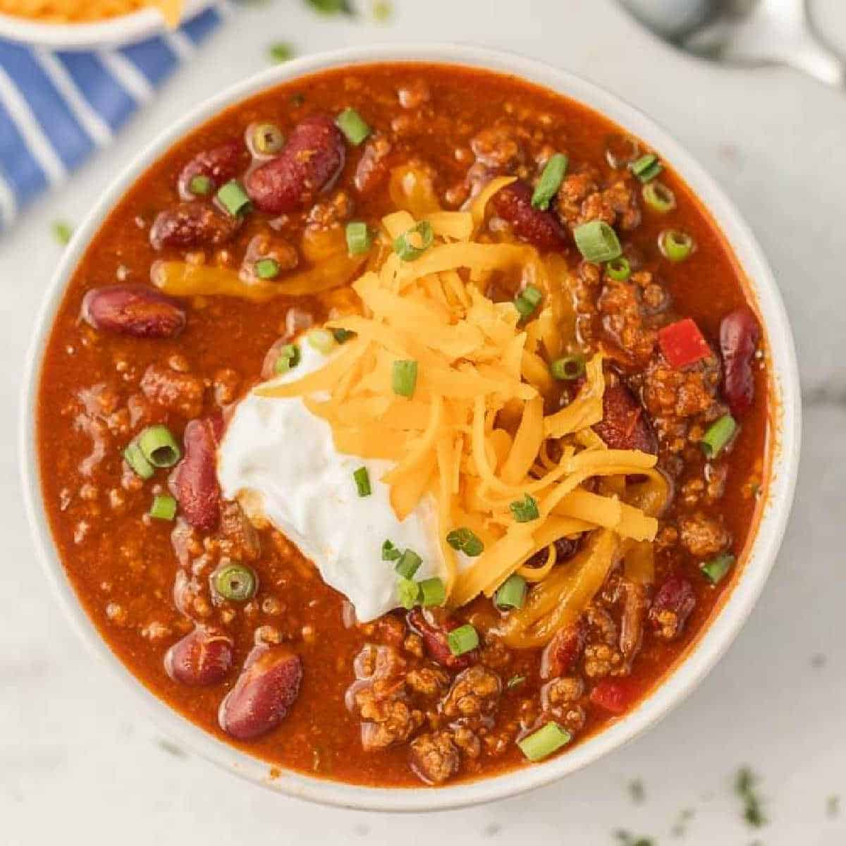 A bowl of Instant Pot Cowboy Chili topped with shredded cheddar cheese, a dollop of sour cream, and chopped green onions, featuring red beans and ground meat in a thick, red sauce.