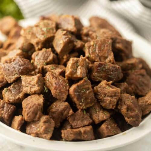 A white plate filled with browned, seasoned beef cubes—an Instant Pot Steak Bites recipe delight. The meat is piled high, showcasing a slightly crispy exterior with hints of green herbs sprinkled on top. A striped cloth is partially visible in the background.