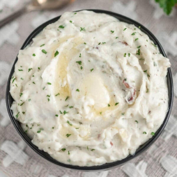 A bowl of creamy Ranch Mashed Potatoes topped with melted butter and sprinkled with chopped parsley, placed on a textured light-colored cloth.