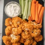 A platter featuring our Roasted Buffalo Cauliflower Recipe, with crispy florets, celery sticks, carrot sticks, and a bowl of ranch dip, all garnished with fresh herbs and arranged on a sleek black tray.