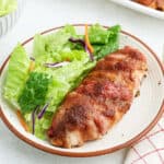 A plate featuring Brown Sugar and Bacon Wrapped Chicken breast, served alongside a leafy green salad with shredded carrots and purple cabbage.