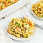 A plate of creamy chicken noodle casserole with carrots and peas is served with a fork. A casserole dish and another plate of the same chicken noodle casserole recipe are in the background on a white surface.