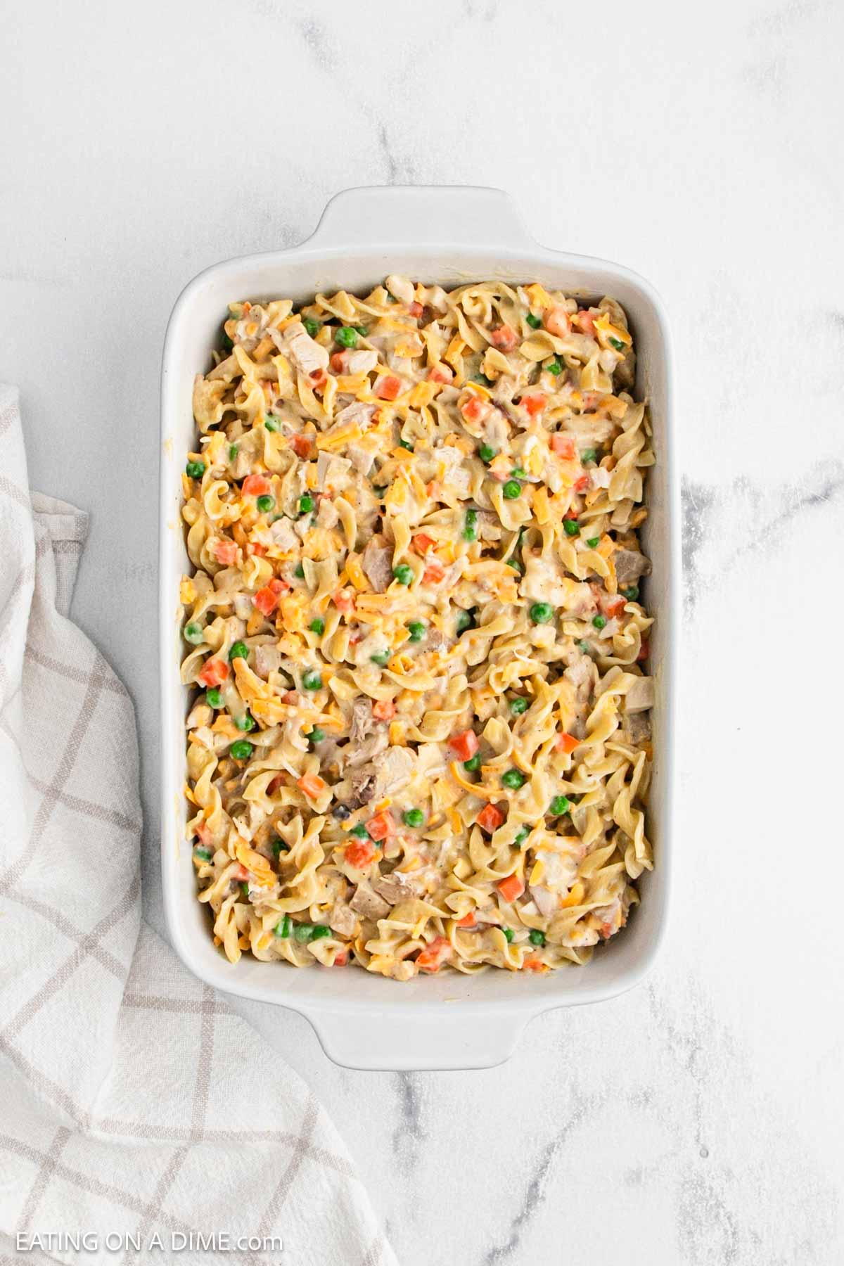 A white casserole dish filled with creamy chicken noodle casserole mixed with peas, carrots, and chunks of meat, placed on a marble surface beside a beige and white checkered towel.