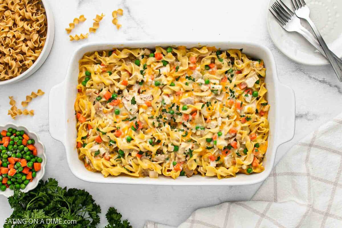 A white baking dish filled with creamy chicken noodle casserole recipe, topped with peas, carrots, and parsley. Bowls of dry noodles and mixed veggies, plates, and forks are arranged nearby on a white marble surface.
