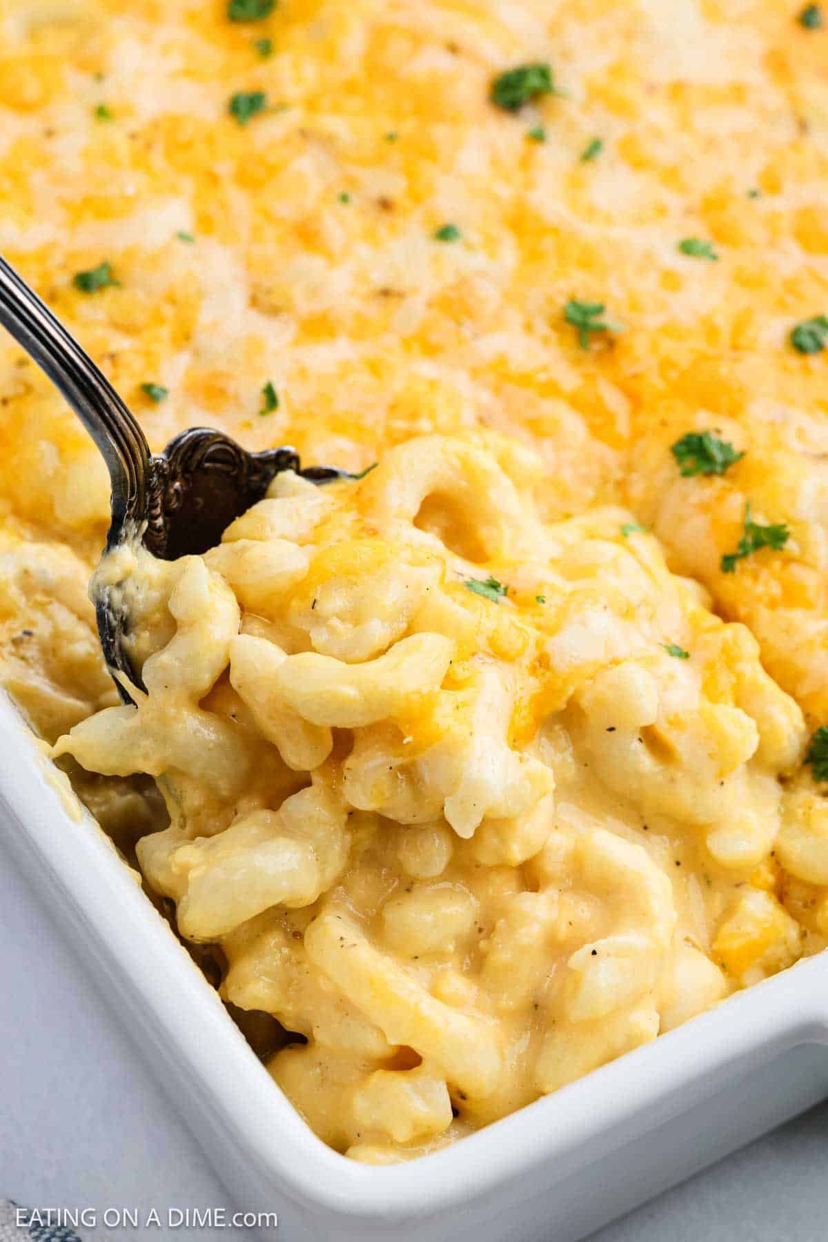 A close-up of creamy Copycat Chick-Fil-A Mac and Cheese in a white dish, with a spoon scooping out a portion. The cheese is melted and golden on top, garnished with a sprinkle of green herbs.