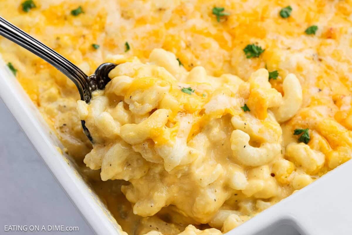 A close-up of creamy Copycat Chick-Fil-A Mac and Cheese in a white dish, with a serving spoon scooping out a portion. The dish is topped with melted cheese and garnished with small bits of parsley.