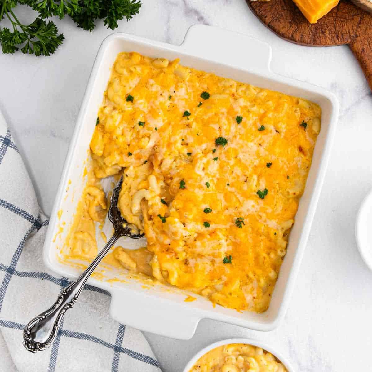 A square white baking dish filled with creamy, baked Copycat Chick-Fil-A Mac and Cheese, topped with melted cheese and parsley, with a serving spoon and some macaroni scooped out.