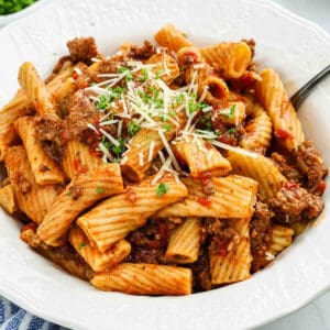 A white bowl filled with rigatoni pasta mixed with ground meat and tomato sauce, inspired by a crock pot Italian sausage rigatoni recipe, is topped with grated cheese and chopped herbs, sitting on a table with a fork.