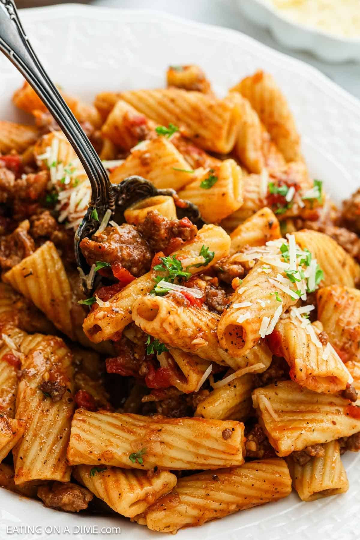 A close-up of rigatoni pasta with ground beef, tomato sauce, and herbs, inspired by a crock pot Italian sausage rigatoni recipe. Grated cheese and chopped parsley top the dish as a fork lifts a portion from the plate.