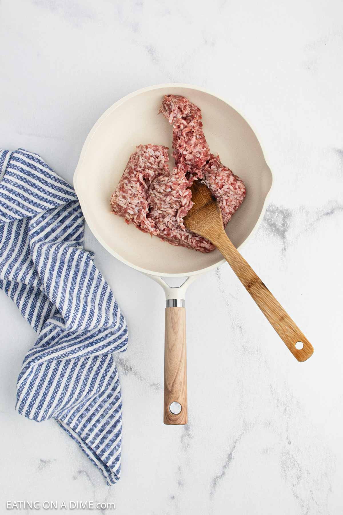 Raw ground beef in a white skillet is being stirred with a wooden spoon, an essential step for any crock pot Italian sausage rigatoni recipe. A blue and white striped kitchen towel lies beside the pan on a white marble countertop.
