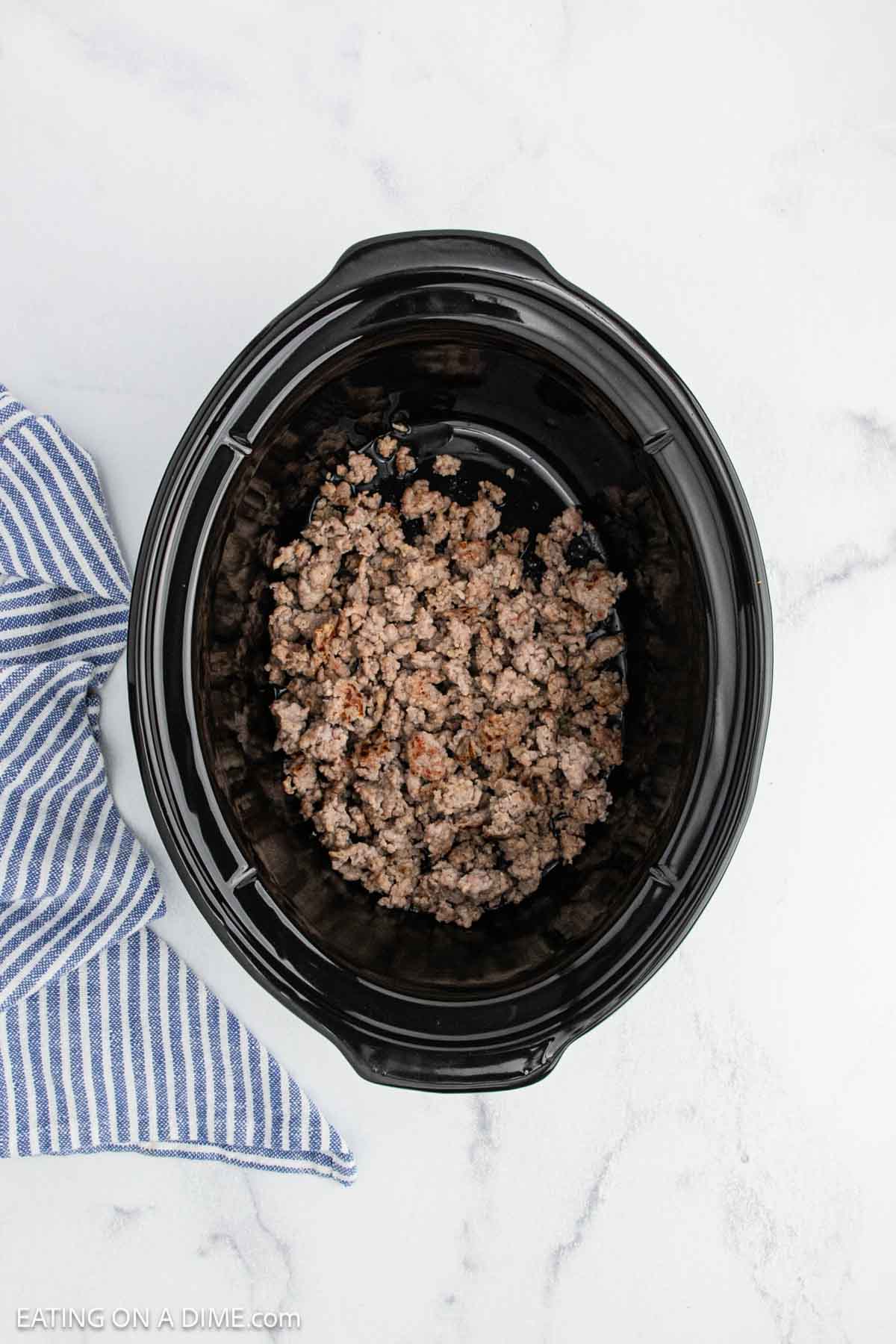A black crock pot containing cooked ground beef sits on a white marble surface next to a blue and white striped kitchen towel, ready for your Italian sausage rigatoni recipe.