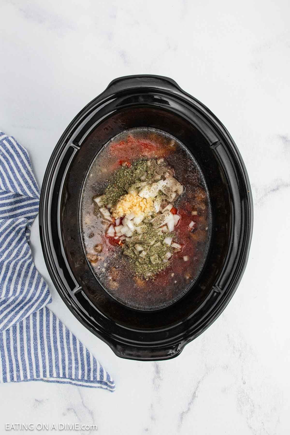A crock pot filled with raw ingredients, including onions, garlic, herbs, and liquids for an Italian sausage rigatoni recipe, sits on a white surface next to a blue and white striped kitchen towel.