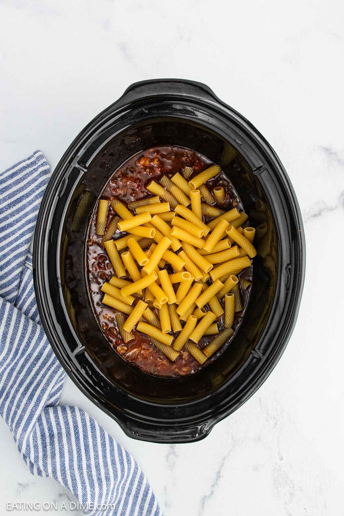 A black crock pot containing uncooked rigatoni pasta atop a savory meat and tomato sauce mixture, part of a delicious crock pot Italian sausage rigatoni recipe, sits on a white marble surface next to a blue and white striped towel.