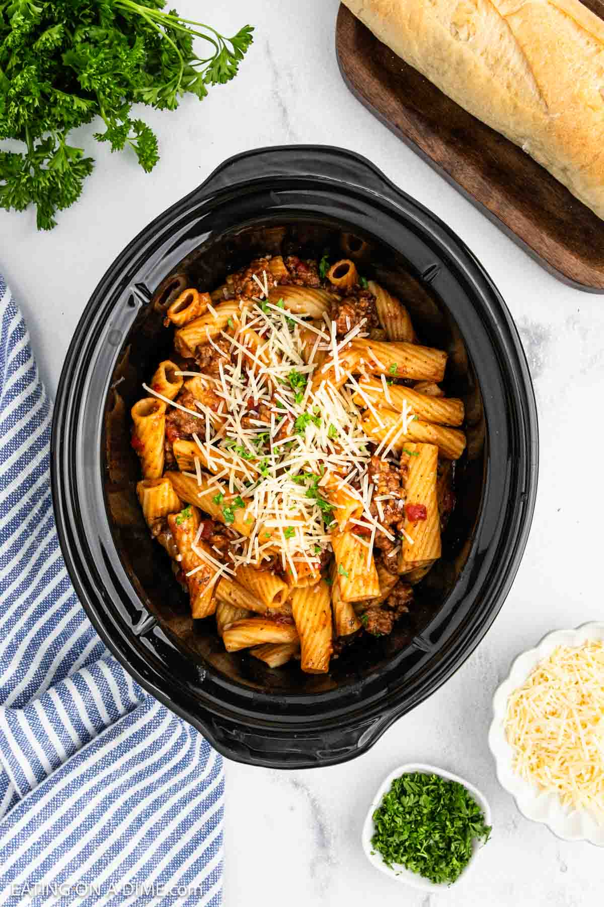 A black slow cooker filled with crock pot Italian sausage rigatoni recipe, topped with shredded cheese and parsley, sits on a white marble surface. Surrounding it are fresh parsley, a loaf of bread, grated cheese, and a blue-striped towel.
