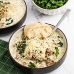 A bowl of creamy instant pot zuppa toscana with ground sausage, chopped kale, potato pieces, and shredded cheese on top, garnished with a piece of bread. A spoon is in the bowl, and a dish of fresh kale is in the background.