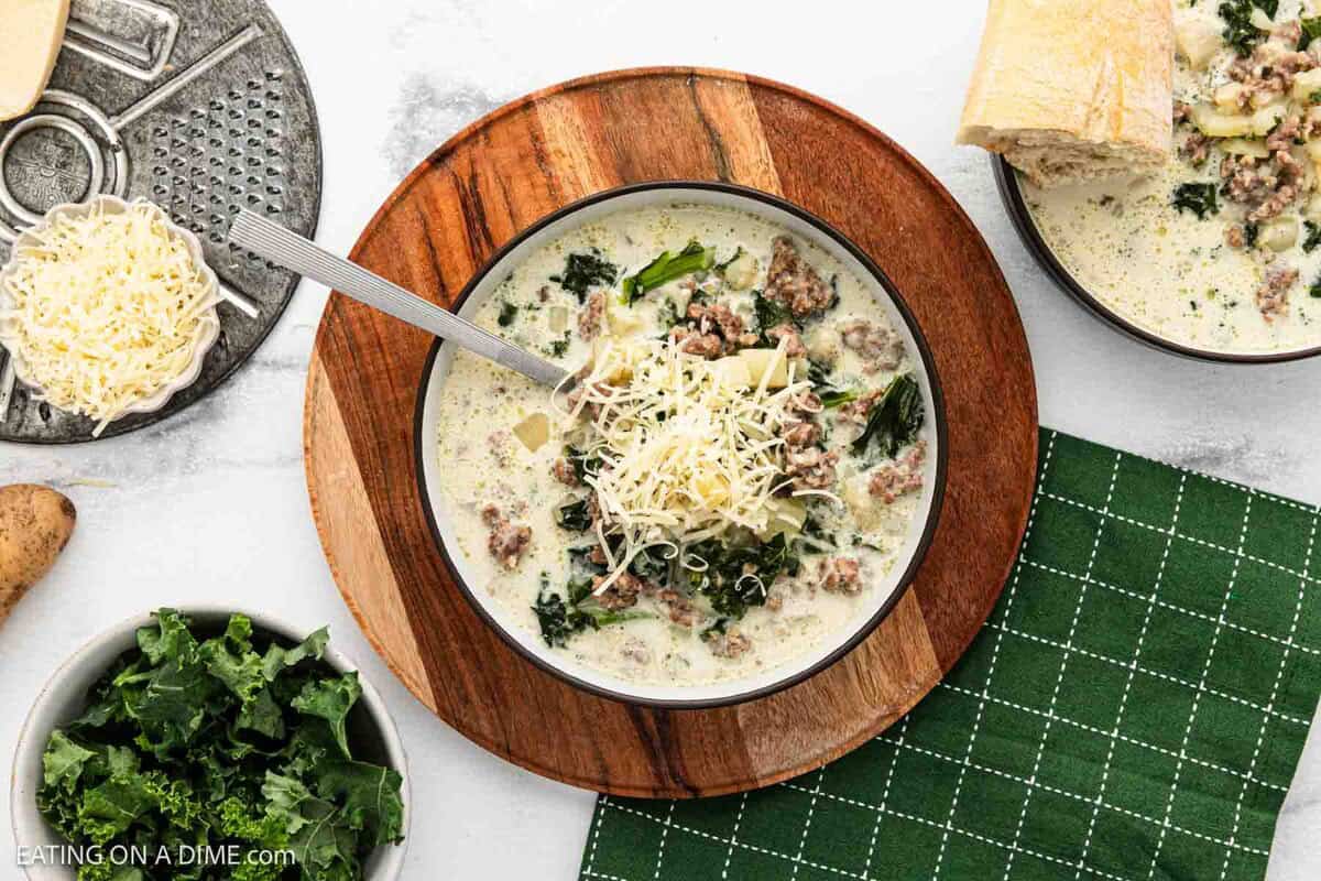 An overhead view of a bowl of creamy sausage and kale soup on a wooden board surrounded by a green napkin, fresh kale, and a bowl of shredded cheese.