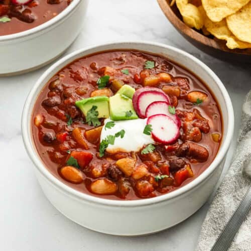 A bowl of Instant Pot vegetarian chili topped with sliced avocado, radishes, sour cream, and cilantro sits on a white surface next to a bowl of corn chips.