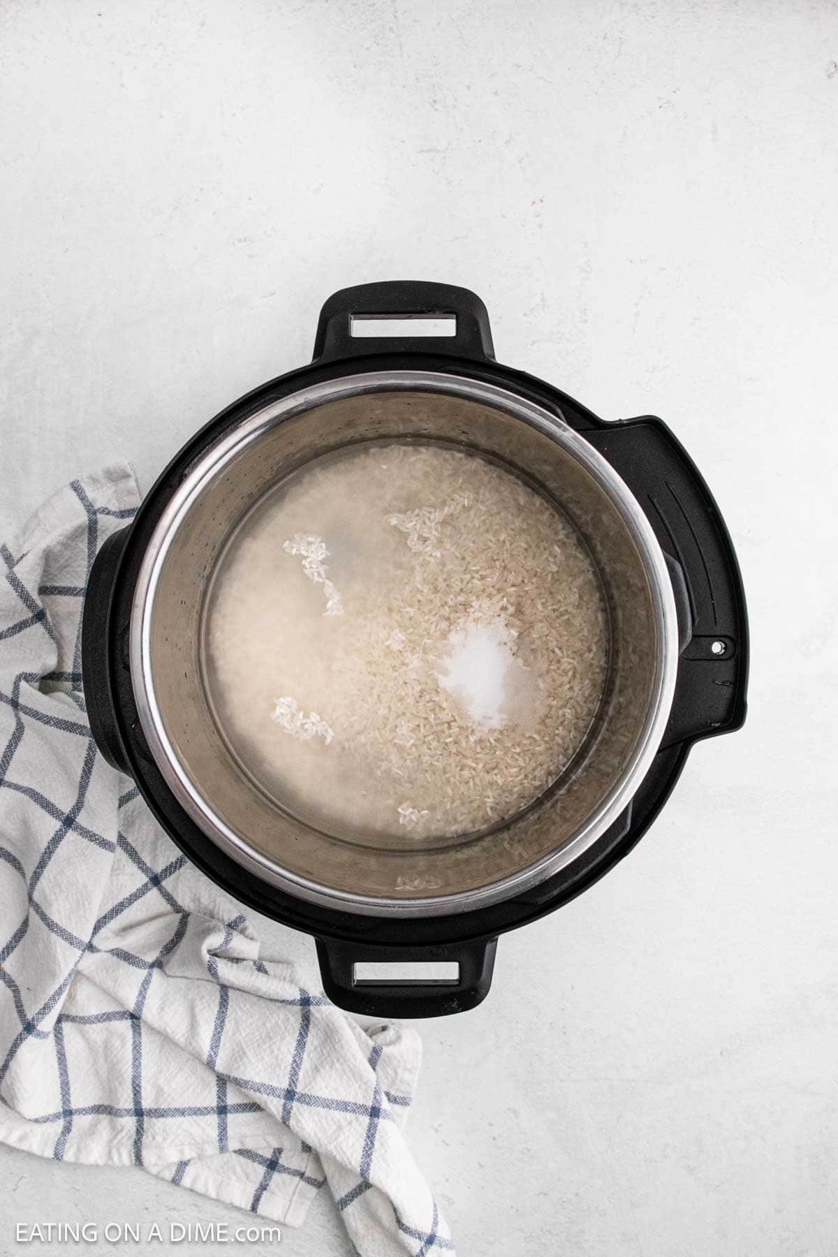 Top-down view of an Instant Pot with uncooked Instant Pot white rice, water, and a bit of salt, set on a light surface alongside a white and blue checkered kitchen towel.