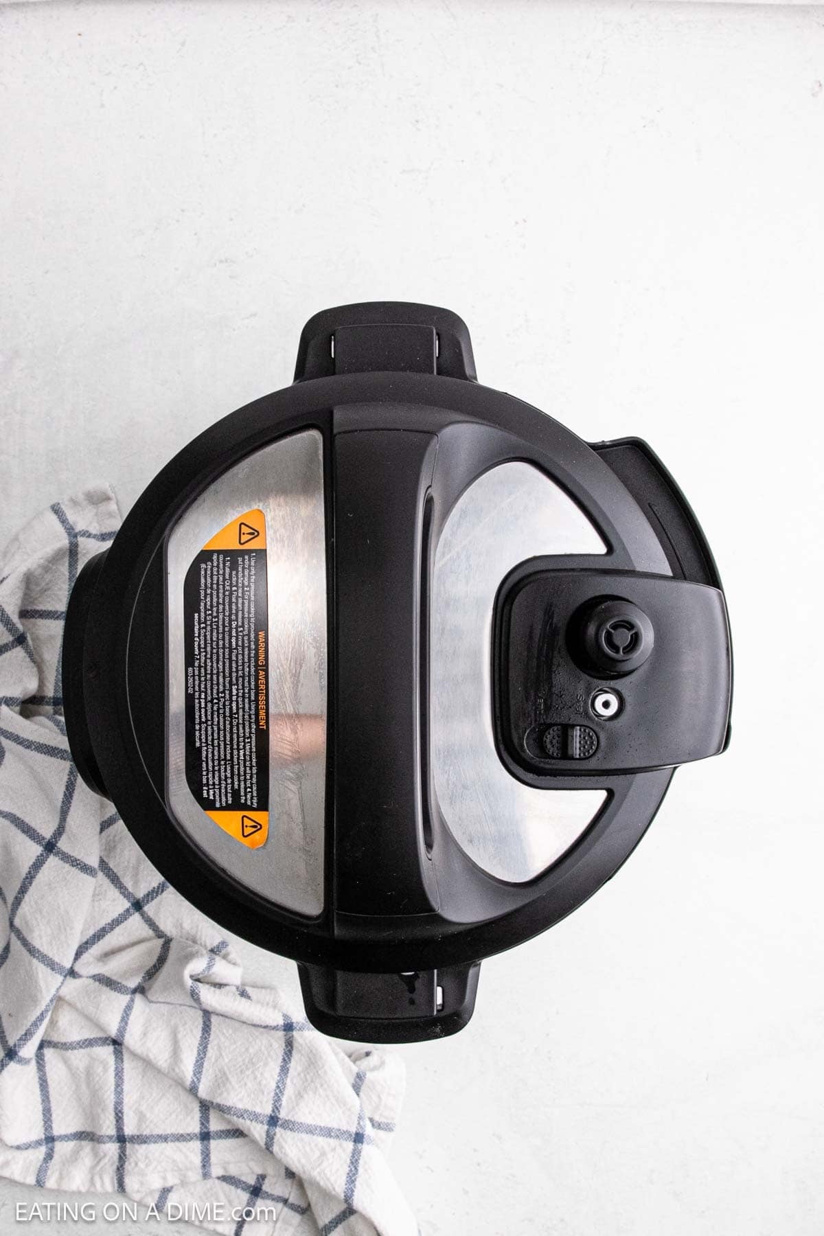 An overhead view of a closed black and silver Instant Pot electric pressure cooker on a white surface, with a blue and white checkered kitchen towel beside it—perfect for making fluffy Instant Pot white rice.
