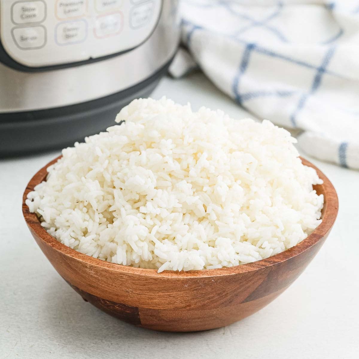 A wooden bowl filled with perfectly cooked Instant Pot white rice sits on a white surface, with a slow cooker and a white-and-blue checkered towel in the background.