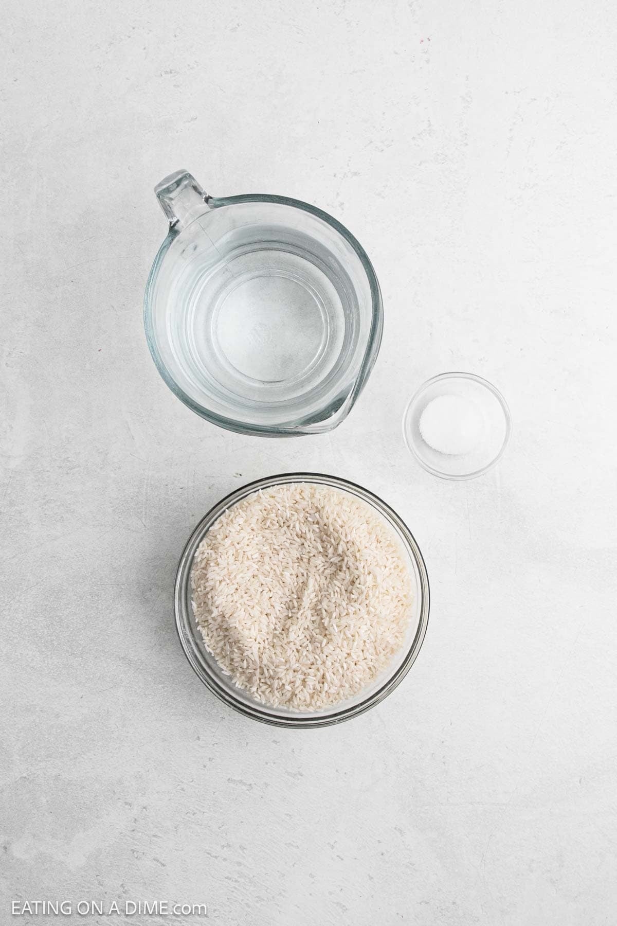 A glass bowl of uncooked rice, a glass measuring cup filled with water, and a small glass bowl of salt are arranged on a light-colored surface—perfect ingredients for making Instant Pot white rice.