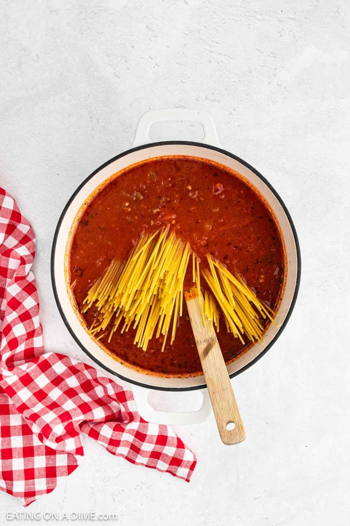 Uncooked spaghetti noodles added to the pasta sauce in a large pot with a wooden spoon.
