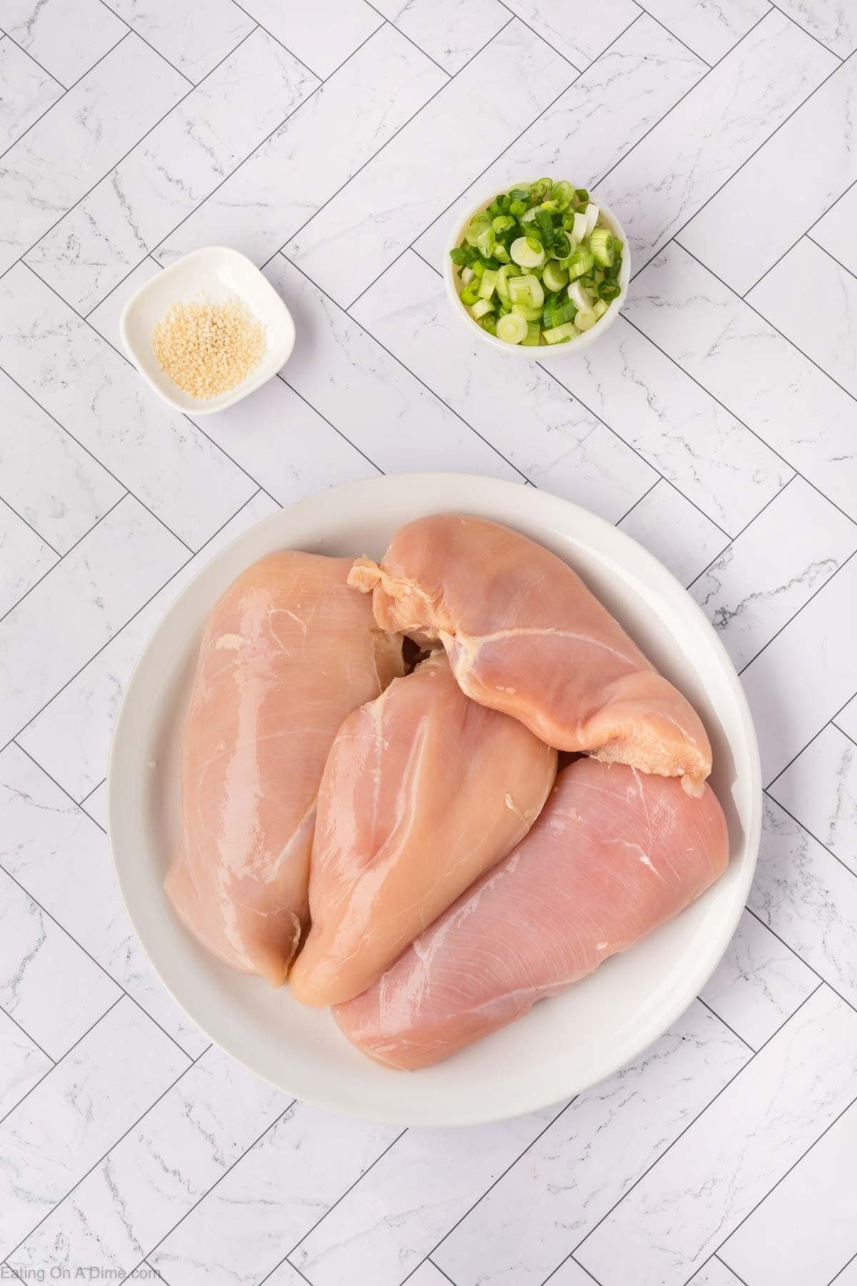 A white plate with raw chicken breasts on it sits on a marble-pattern counter, ready for a baked teriyaki chicken recipe. Nearby are two small bowls, one filled with chopped green onions and the other with white sesame seeds.