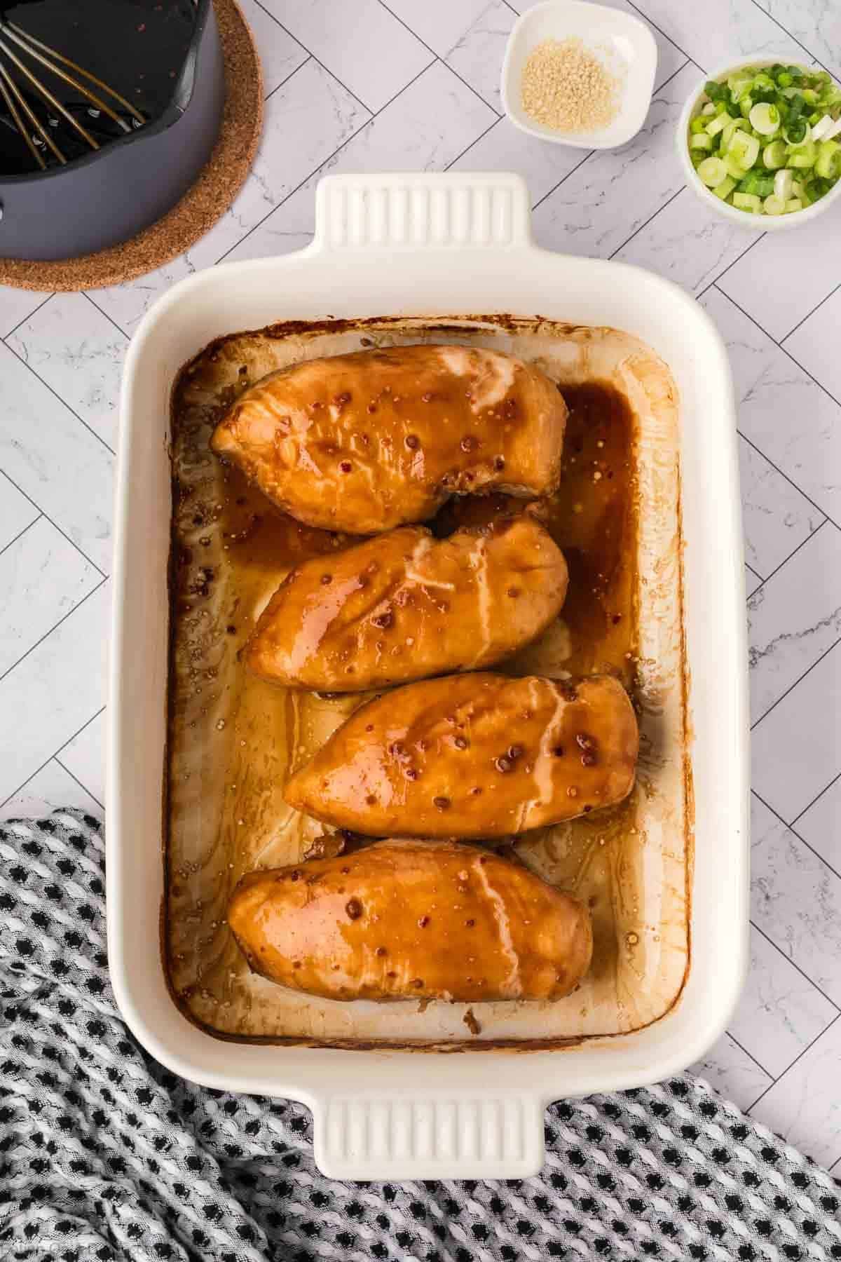 Four glazed chicken breasts in a white baking dish, sitting on a patterned towel. Small bowls of sesame seeds and chopped green onions are nearby—perfect for serving with this baked teriyaki chicken recipe.
